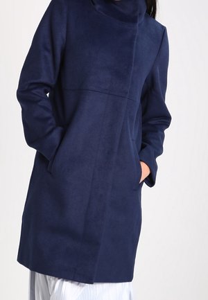 Femme portant un manteau bleu marine mi-longues manches longues, les mains dans les poches, associé à une jupe plissée blanche.