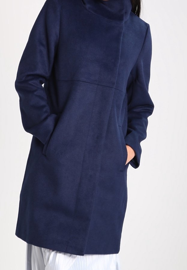 Femme portant un manteau bleu marine mi-longues manches longues, les mains dans les poches, associé à une jupe plissée blanche.