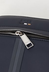 Marine lærveske med glidelås, som har sølvfarget hardware, en teksturert stripe, og preget logo i svart, hvitt og brunt.