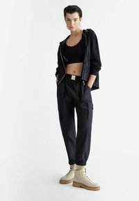 Giacca nera corta con zip e texture a righe, top nero senza maniche, pantaloni cargo neri larghi, stivaletti beige con lacci e platform.