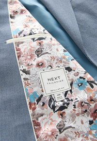 Intérieur de veste de costume avec doublure à motifs floraux dans des couleurs pastel, comportant une étiquette "NEXT Tailoring Regular Fit" et une taille 40R.