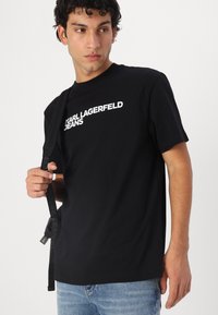 Juodas medvilnės marškinėliai trumpomis rankovėmis su baltu grafiniu logotipu "KARL LAGERFELD JEANS" per krūtinę. Dėvimi su mėlynomis džinsinėmis kelnėmis.