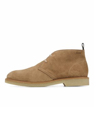 Bottes ankle en suede tan avec deux œillets, semelle en crêpe, anse noire et logo "BOSS" embossé près du talon.