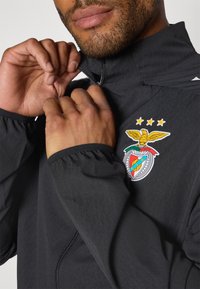 Sort lynlåsejakke med høj krave, prydet med et farverigt emblem med en ørn og stjerner på venstre side. Tekstureret stof, elastiske manchetter.