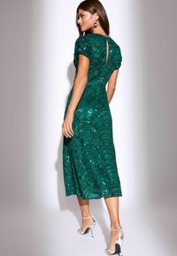 Emeraldzaļš kleita ar sekvīru viļņu rakstu, īsām piedurknēm, atslēgas formas izgriezums aizmugurē, līdz ceļgaliem, saskaņota ar sudraba augstpapēžu sandālēm.