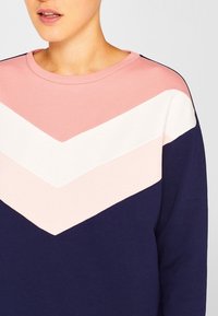 Sudadera de manga larga en color marino, con un patrón de chevrón que presenta secciones en rosa claro, blanco y rosa pálido. Cuello redondo, material suave.