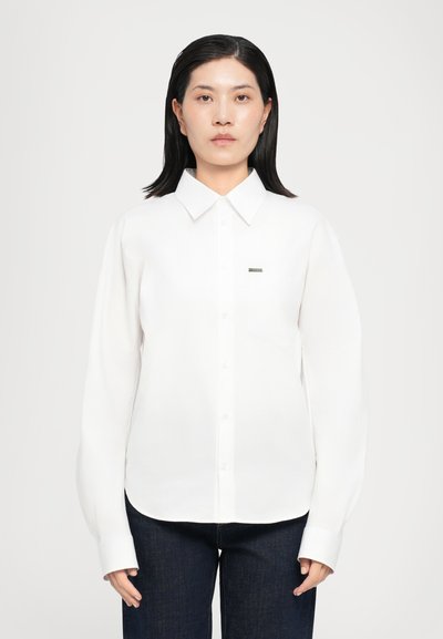 Chemise blanche à manches longues avec un col structuré, poche poitrine discrète et logo brodé. Texture lisse et coupe ajustée.