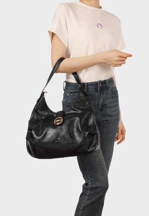 Donna che indossa una maglietta a maniche corte color avorio e jeans scuri, con una grande borsa a tracolla in pelle nera con chiusura dorata.