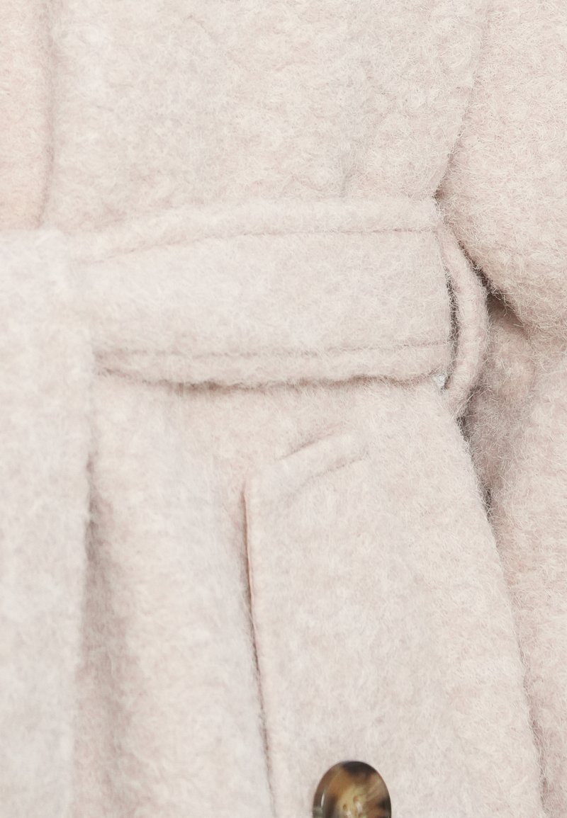 Cappotto di lana rosa chiaro con finitura testurizzata, dotato di una cintura, chiusura con grandi bottoni e tasche laterali. Aspetto morbido e accogliente.