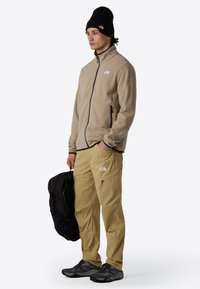 Beige fleece jacket with black zipper, khaki cargo pants with logo, black beanie, holding a black backpack, and gray hiking shoes.
