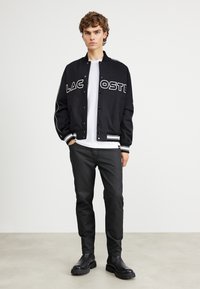 Svart varsityjacka med "LACOSTE"-logotyp, vita detaljer, ribbade ärmar, bärs över en vit T-shirt, i kombination med svarta byxor och stövlar.