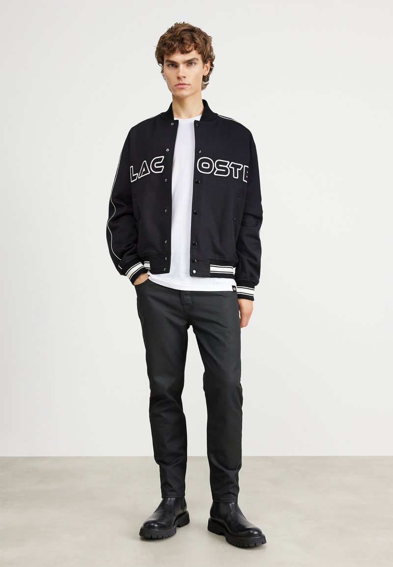 Veste de varsity noire avec logo "LACOSTE", accents blancs, poignets côtelés, portée sur un t-shirt blanc, accompagnée de pantalons noirs et de bottes.