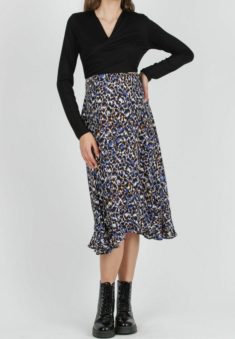 Femme portant un haut cache-cœur noir à manches longues, une jupe midi imprimé animalier bleu et marron, et des bottes noires à lacets, debout avec une main sur la hanche.