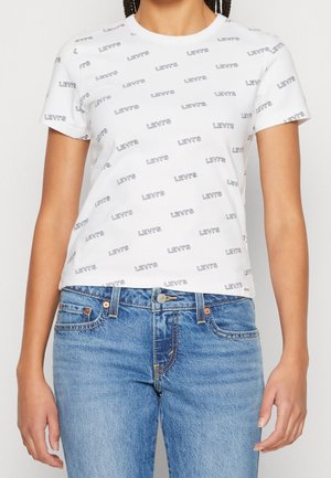 T-shirt blanc à manches courtes avec motif de texte noir répétée "Levi's", porté avec un jean bleu sur une personne debout.