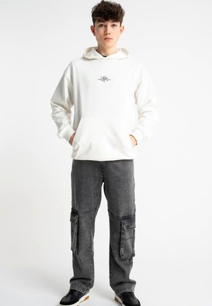Nastolatek w białej bluzie z kapturem z małym logo na piersi, szarych spodniach bojówkach i czarnych trampkach, stojący na białym tle.