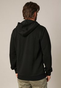 Haut à capuche noir texturé avec un motif en grille, doté d'une capuche, de poignets et d'un ourlet côtelés, et d'une coupe décontractée, vu de dos.