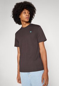 Mørkebrun t-skjorte i bomull med korte ermer, rund hals og en liten turkisbla logo på venstre bryst. Brukt sammen med lyseblå jeans.