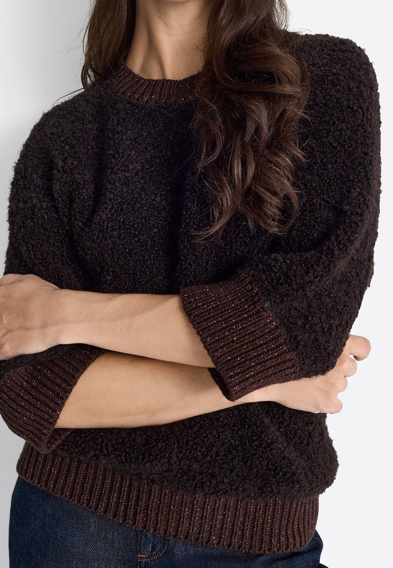 Personne aux longs cheveux bruns ondulés portant un pull texturé marron foncé et un jean bleu, les bras croisés devant le torse.