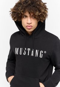 Felpa nera in cotone con un grande logo bianco "MUSTANG". Presenta un cappuccio con coulisse e una tasca a marsupio frontale. Tessuto morbido.