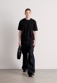 Fekete rövid ujjú póló, sötét, bővülő farmerral párosítva. A modell egy fekete bőrtáskát visel, semleges háttér előtt sétálva.