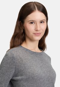 Junge Frau mit schulterlangem braunem Haar, trägt einen grauen Strickpullover, blickt nach vorne mit einem leichten Lächeln vor einem weißen Hintergrund.