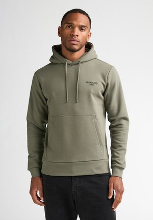 Mann trägt ein olivgrünes Kapuzensweatshirt mit Fronttasche und schwarze Hose, steht vor einem einfarbigen weißen Hintergrund.
