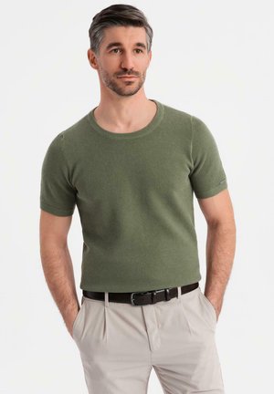 Suéter de punto de manga corta en verde oliva con cuello redondo, de tejido texturizado, combinado con pantalones beige claros y un cinturón marrón.