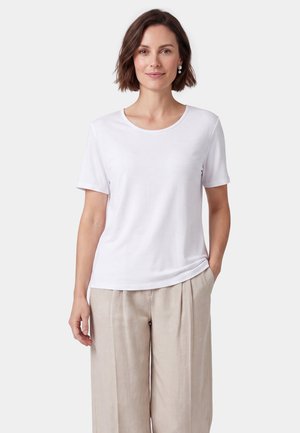 Donna con capelli corti castani che indossa una camicia bianca a maniche corte e pantaloni beige a gamba larga, in piedi con una mano in tasca su sfondo bianco.