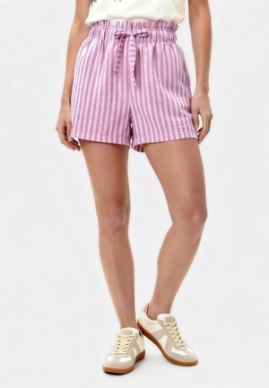 Kvinde iført pink og lys lilla stribede shorts med knyttet taljebånd og beige sneakers på hvid baggrund.
