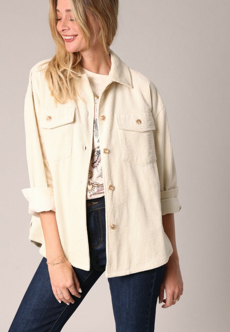 Veste en velours côtelé crème avec un col, poches poitrine et fermetures à boutons. Manches retroussées et coupe décontractée.