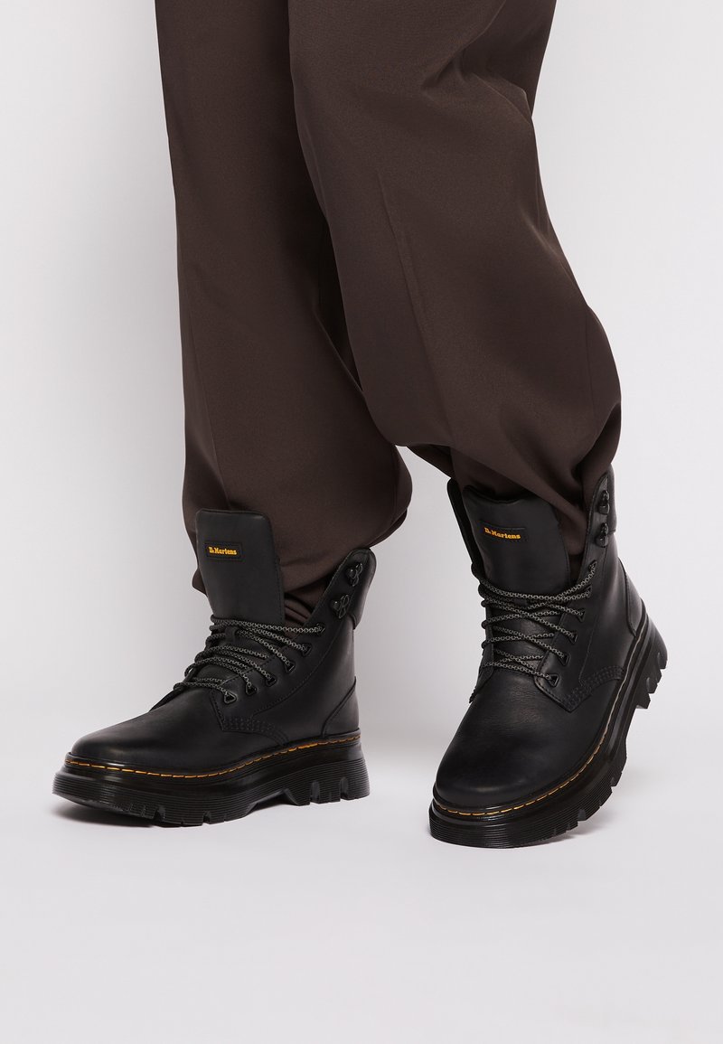 Bottes noires en cuir à lacets avec semelles épaisses, portées avec un pantalon ample marron foncé sur un fond clair uni.