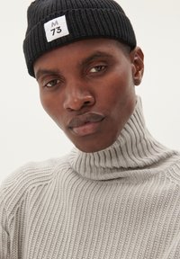 Pulover gri cu guler înalt și textură ripată, cu un design ajustat. Este purtat cu o căciulă neagră din tricot, care are o etichetă albă inscripționată cu "M 73."