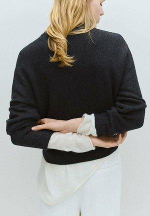 Jersey de lana gris oscuro con un ajuste relajado y hombros caídos, superpuesto a una camisa blanca texturizada, mostrando colores y materiales en contraste.