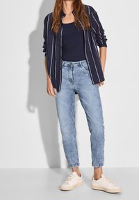 Navy striped button-up shirt over a dark tank top, light blue acid-wash jeans, and beige sneakers. Casual outfit with relaxed fit.