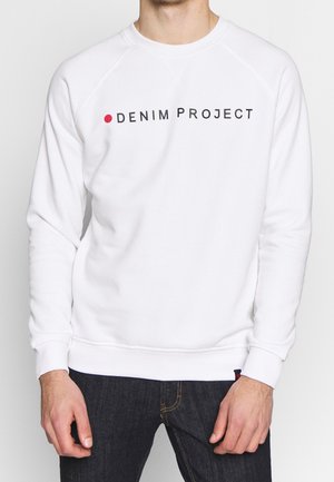 Mand iført hvid sweatshirt med teksten "Denim Project" og en rød prik på brystet, kombineret med mørkeblå jeans.