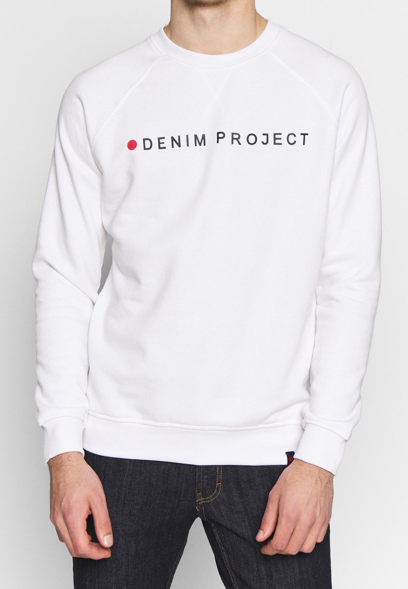 Homme portant un sweat-shirt blanc avec le texte « Denim Project » et un point rouge sur la poitrine, associé à un jean bleu foncé.