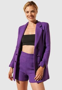 Blazer violet texturé avec short assorti. Le blazer présente des boutons croisés à double boutonnage ; le short est ajusté et cintré. Crop top noir en dessous.