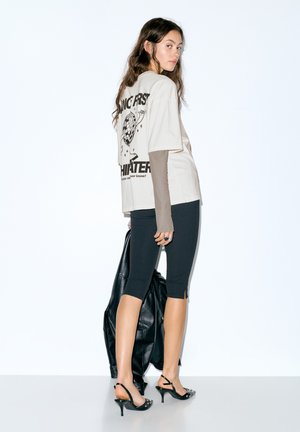 Kremna grafična majica s črnim napisom in obliko, nošena čez rjavo majico z dolgimi rokavi. Črna jakna iz umetnega usnja, črne legice in salonarji.