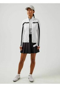 Giacca bianca con zip e dettagli neri, abbinata a una gonna nera plissettata. Il modello indossa un cappellino grigio chiaro e scarpe da ginnastica bianche.