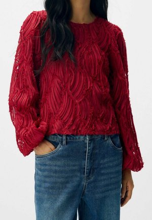 Femme portant un pull rouge texturé à manches longues avec un motif en relief et un jean bleu, une main dans la poche, sur fond blanc.