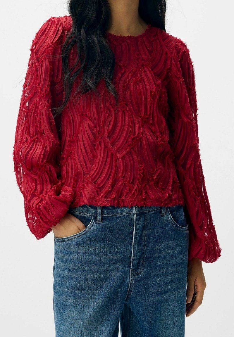 Femme portant un pull rouge texturé à manches longues avec un motif en relief et un jean bleu, une main dans la poche, sur fond blanc.