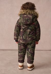 Kamuflāžas snowsuit zaļā un brūnā krāsā ar pūkainu kapuci, elastīgām aprocēm un brūnām kurpēm ar mākslīgo kažoku. Sarkans logotipa detalizējums.