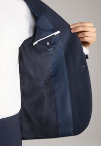 Interno di una giacca da uomo blu con una fodera in satin liscia e un motivo geometrico, etichettata taglia 8, e un accento bianco vicino alla tasca.