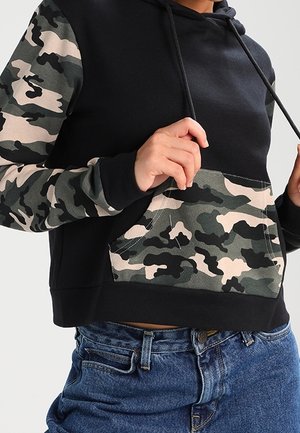 Personne portant un sweat à capuche noir avec des manches et une poche camouflage vert et beige, associé à un jean bleu.