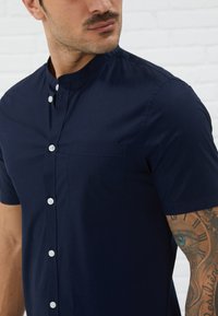 Navy short-sleeve shirt in lightweight fabric featuring a standing collar, front buttons, and a chest pocket. Smooth texture with subtle sheen.