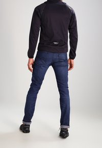 Sort letvægtsjakke med lynlåset krave, tætsiddende ærmer og reflekterende detaljer, parret med mørkeblå jeans og sorte sneakers.