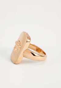 Bague chevalière rectangulaire dorée avec un emblème en relief de tête de Méduse et des détails décoratifs le long du bord supérieur.