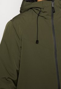 Giacca verde oliva con una texture liscia, cappuccio regolabile e zip nera. Presenta un design geometrico delle cuciture e dettagli con cordini.
