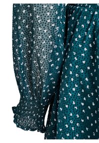 Tirkizna bluzica s uzorkom bijelih i crnih točkica, s pufastim rukavima i sakupljenim zapešćem. Poluprozirna tkanina s laganom, lepršavom teksturom.