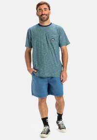 Juostuota trumpo rankovė t-shirt, mėlyna ir tamsiai mėlyna, su priekiu kišenėje su logotipu; derinama su mėlynu šortų ir juodais sportbačiais.